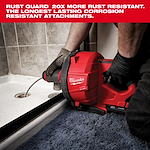 A worker uses a Milwaukee FUEL drain cleaner to clear a shower drain. The machine is red and has an attached cable. Black, gloved hands hold the device steady while the machine works on the drain. Rugged tiles and a blue carpet surround the shower area.