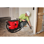 A worker uses a Milwaukee M18 cordless wet/dry vacuum to clean a construction site. The vacuum is red with black accents and has a long hose. The worker wears a lime green shirt, blue jeans, brown shoes, and a white hard hat.