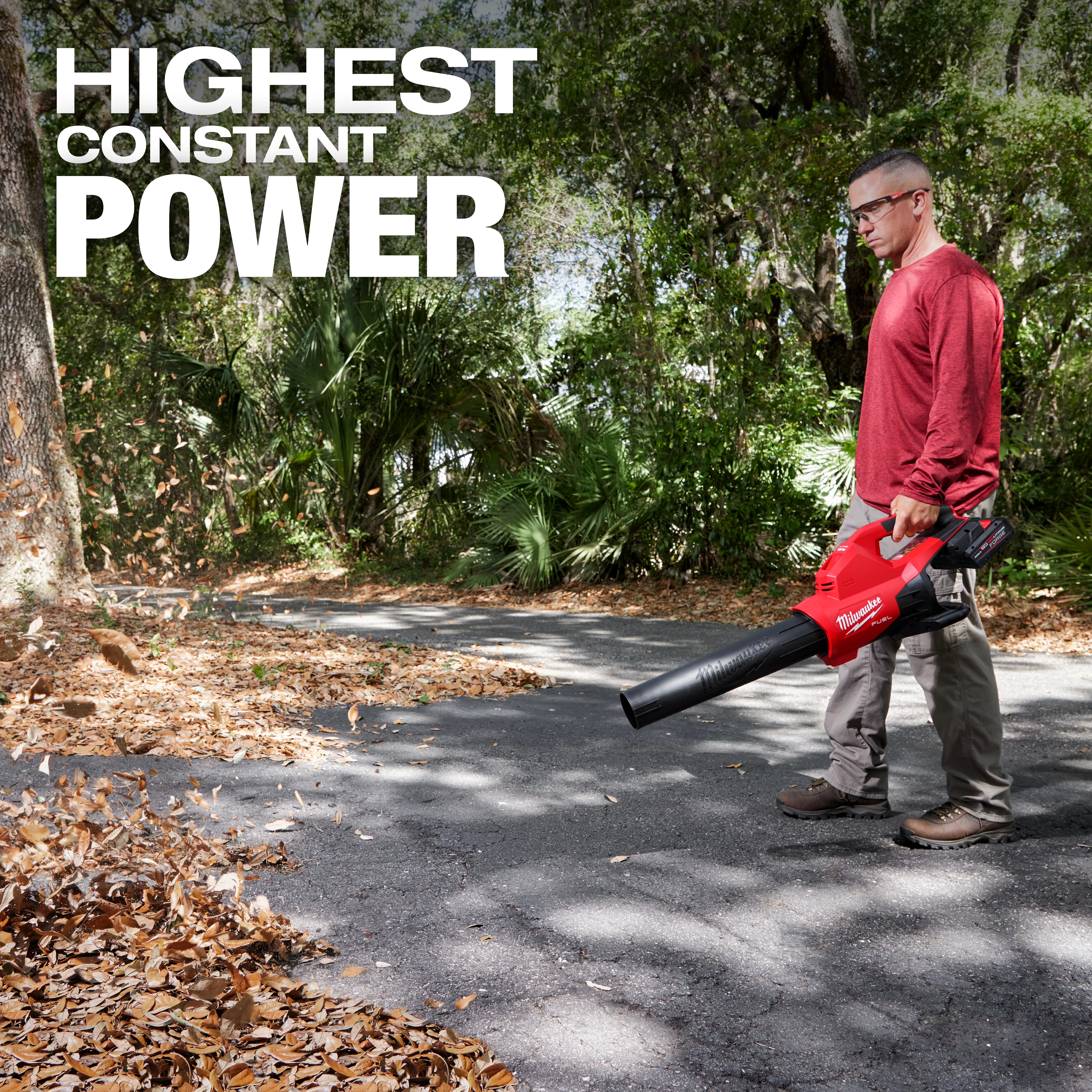 A person is using an M18 FUEL™ Dual Battery Blower to clear leaves from a paved path surrounded by trees. The text "HIGHEST CONSTANT POWER" is displayed at the top left of the image.