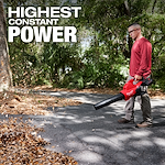 A person is using an M18 FUEL™ Dual Battery Blower to clear leaves from a paved path surrounded by trees. The text "HIGHEST CONSTANT POWER" is displayed at the top left of the image.