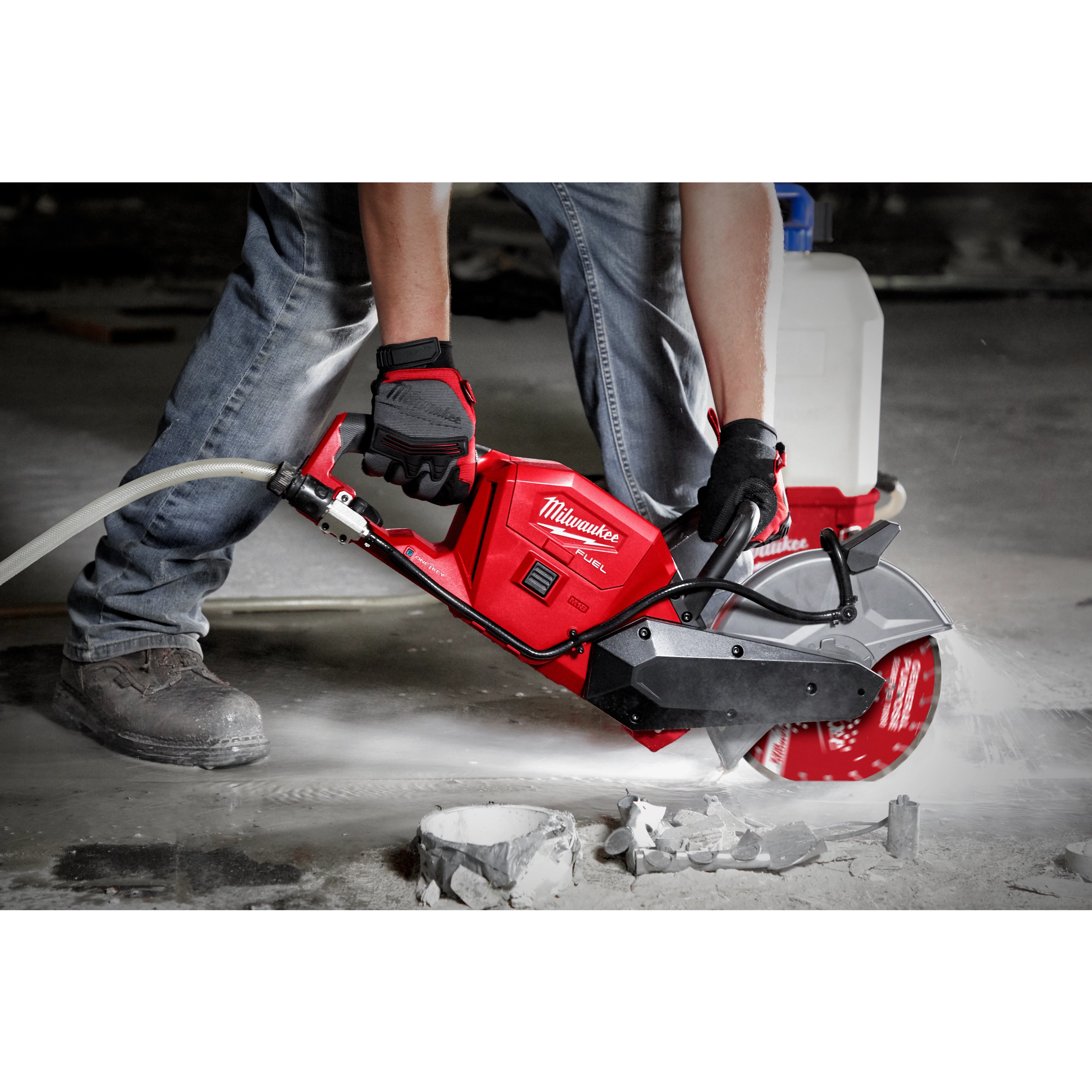 A person wearing gloves operates a red Milwaukee cutting tool to saw through a concrete surface. They wear jeans and sturdy boots. The tool is connected to a water tank, which is visible in the background, to reduce dust. Debris and dust are scattered around the work area.