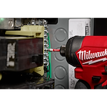 A Milwaukee power tool is being used with a SHOCKWAVE™ 2” Impact Square Recess #2 Power Bit to tighten screws on an electrical panel. The bit is attached to the power tool and is making contact with the screw head. Various wiring components are visible in the background.