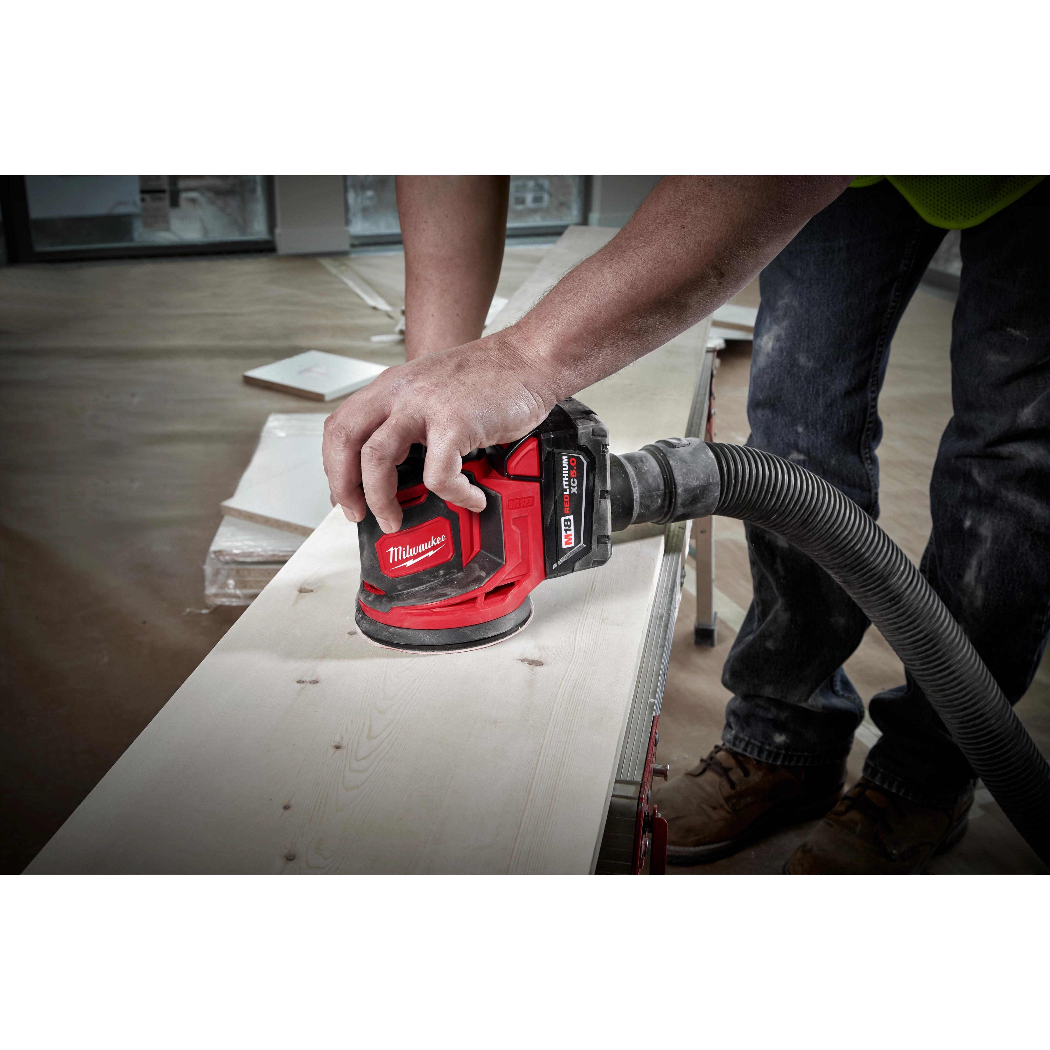 A person using the M18 Random Orbit Sander on a wooden surface. The sander is red and black with a dust extraction hose attached. The person is wearing jeans and work boots. The background shows a spacious, unfinished indoor area with wooden planks and tools.