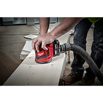 A person using the M18 Random Orbit Sander on a wooden surface. The sander is red and black with a dust extraction hose attached. The person is wearing jeans and work boots. The background shows a spacious, unfinished indoor area with wooden planks and tools.