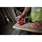 A person sands a wooden plank using an M18 Random Orbit Sander. The worker wears a reflective vest in a construction setting.