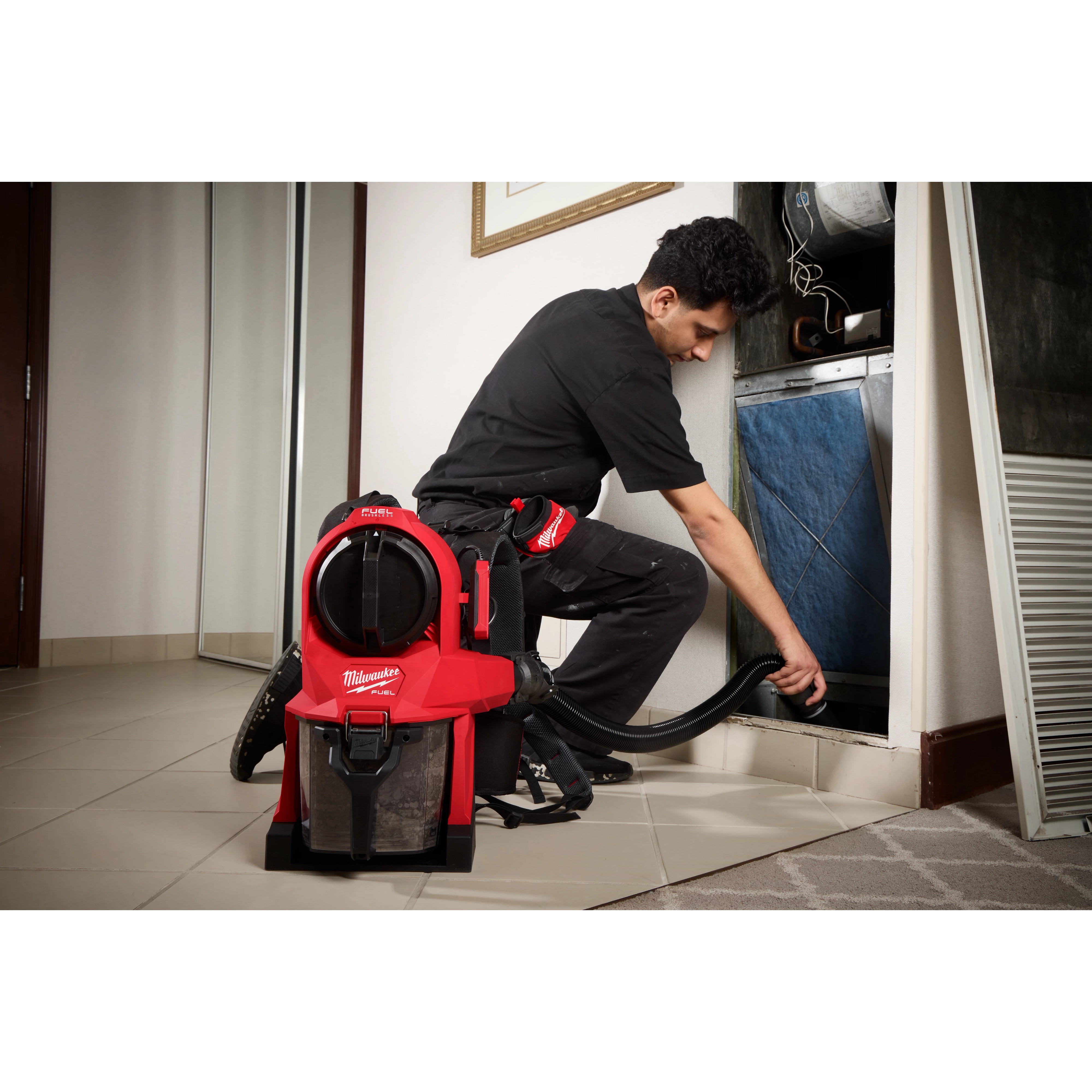 A worker uses a M18 FUEL™ 3-in-1 Backpack Vacuum to clean an HVAC system. The red vacuum is worn on the worker's back and has a black hose attached, ensuring efficient dust removal. The worker wears protective black clothing and kneels beside the HVAC unit.