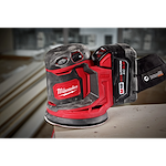 The Milwaukee M18 Random Orbit Sander features ergonomic design with red and black casing, a dust collection system, and a battery pack labeled "M18 REDLITHIUM XC5.0." It is placed on a wooden surface, with building materials visible in the blurred background.