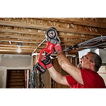 A person uses the M18 FUEL™ Compact Threader with One-Key™ to work on pipes installed in a ceiling. The tool is red and black, and the setting is an unfinished basement or construction site.