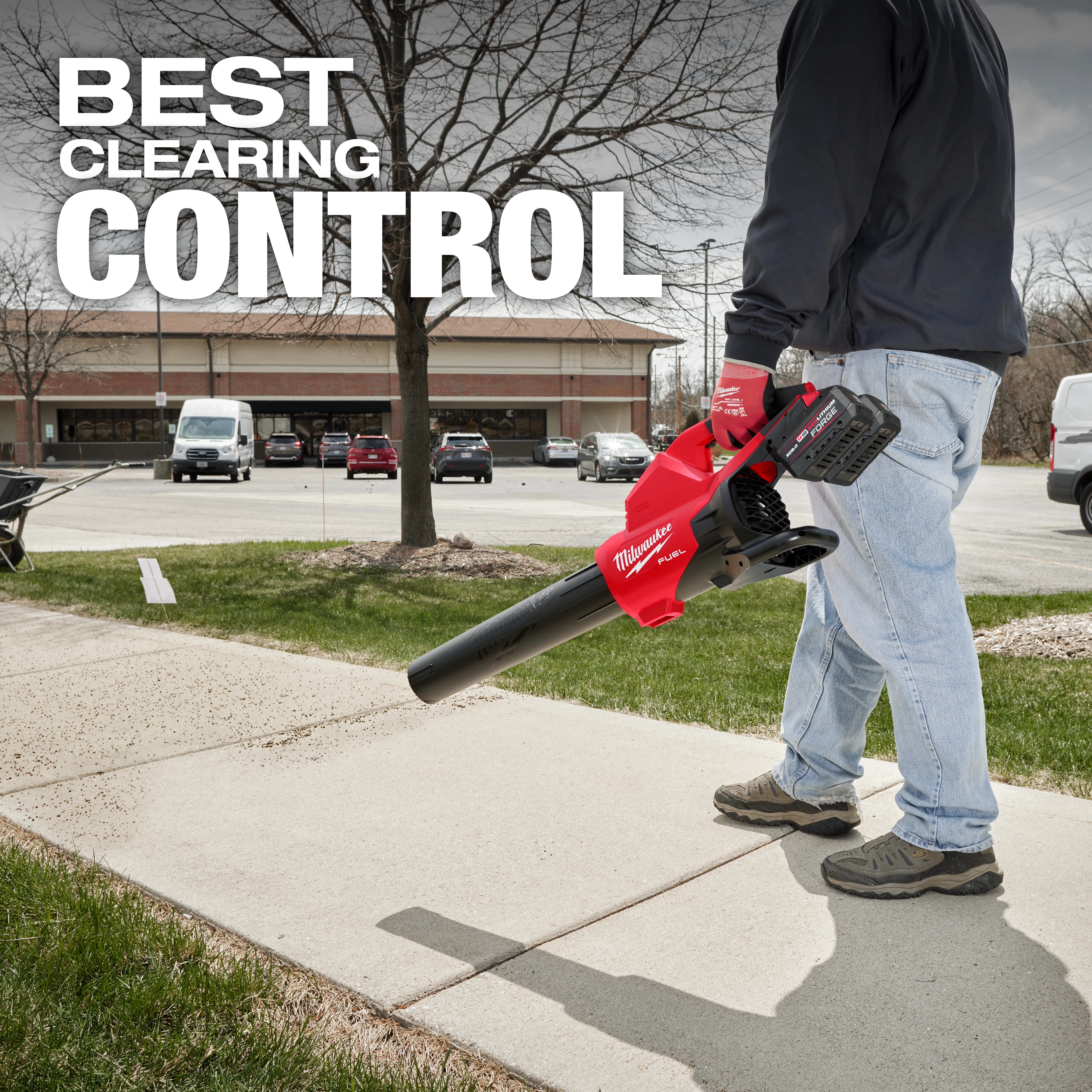 A person uses the M18 FUEL™ Dual Battery Blower to clear a sidewalk. The blower is red and black. The background shows a parking lot with cars, trees, and a building. The text "BEST CLEARING CONTROL" is displayed at the top.
