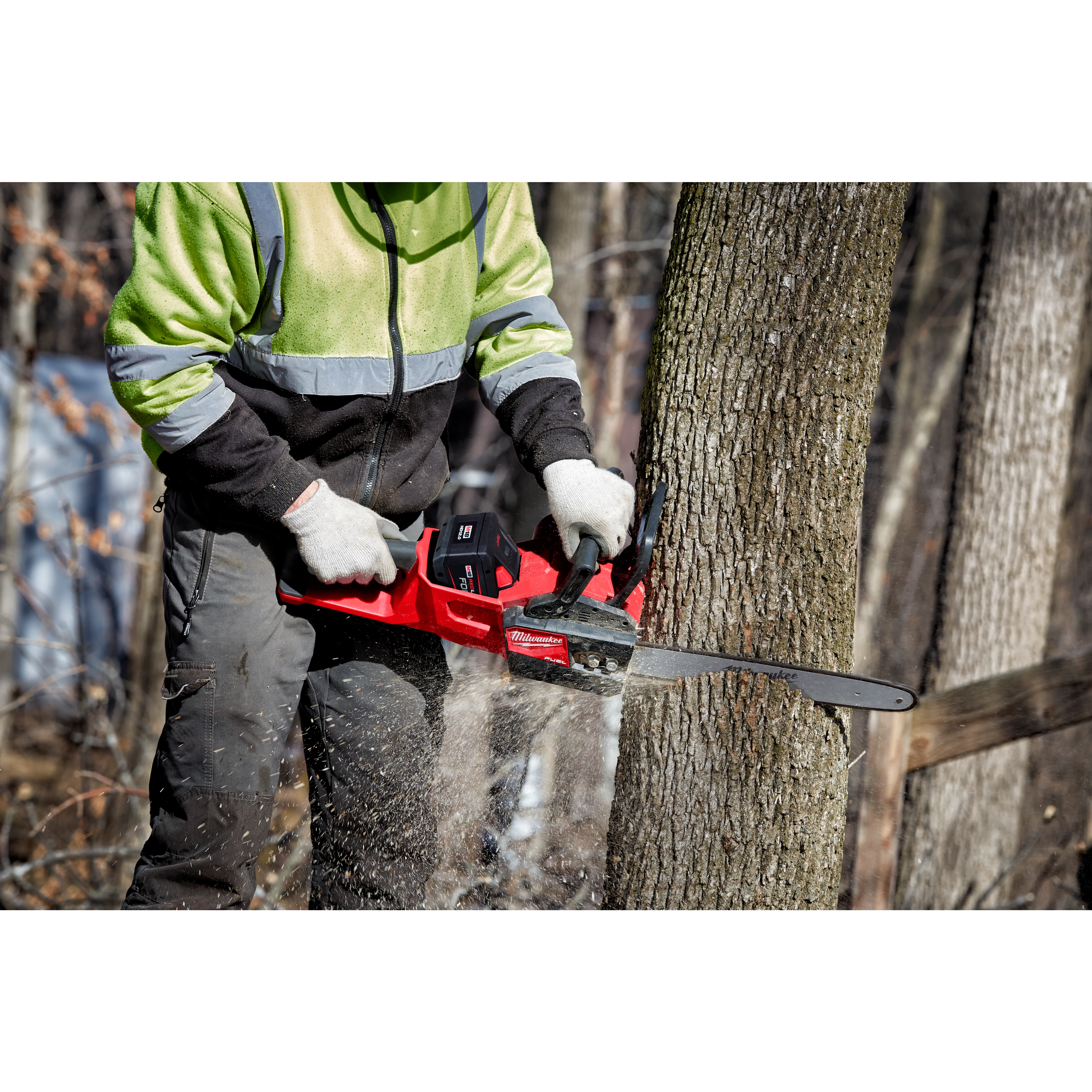 Person using a red chainsaw to cut down a tree trunk in a wooded area, wearing gloves and safety clothing.