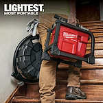 A person carries a Milwaukee Fuel portable air compressor up a wooden staircase. The product slogan "Lightest, Most Portable" appears in bold text at the top left of the image. The compressor is red with black casing and features ergonomic handles for easy transport.