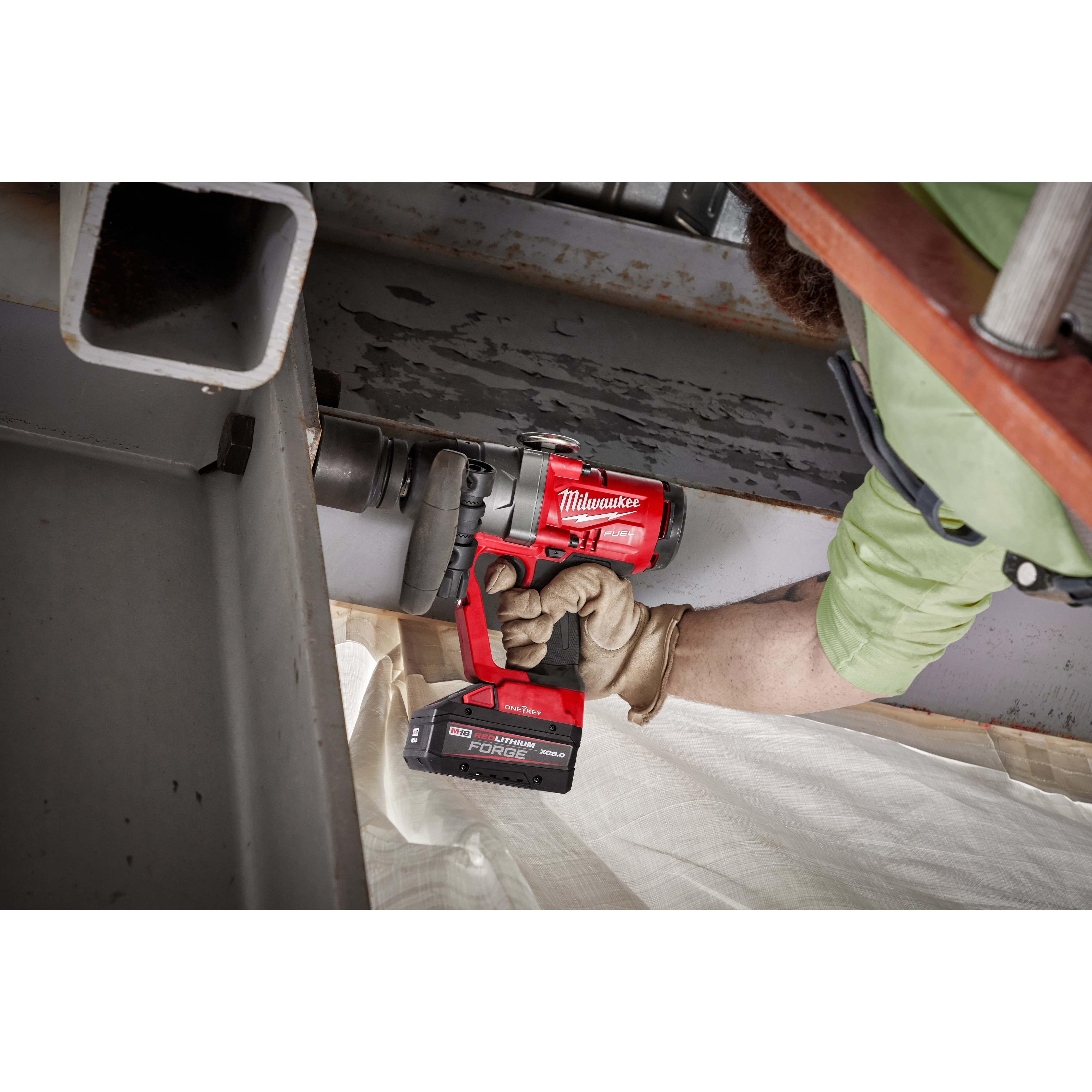 A person in work gloves is using a red M18 FUEL w/ ONE-KEY 1" High Torque Impact Wrench to work on a metal structure. The tool is branded Milwaukee and equipped with a large battery pack.