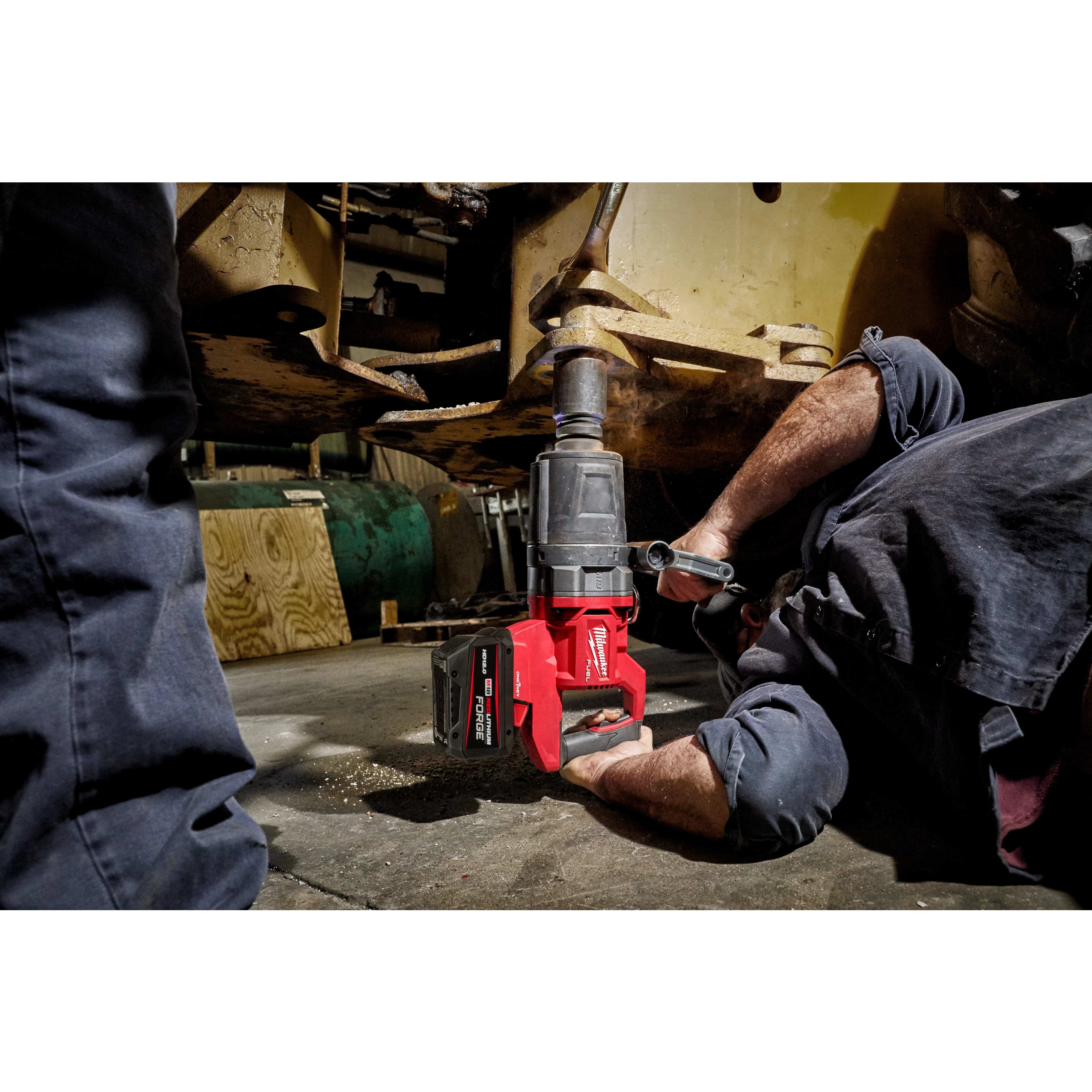 A person operates an M18 FUEL™ 1" D-Handle High Torque Impact Wrench w/ONE-KEY™ App Shot, working on the underside of a large machine. The red and black tool is in use on a bolt or attachment point, highlighting its industrial application.