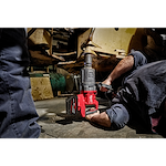 A person operates an M18 FUEL™ 1" D-Handle High Torque Impact Wrench w/ONE-KEY™ App Shot, working on the underside of a large machine. The red and black tool is in use on a bolt or attachment point, highlighting its industrial application.