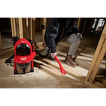 A person uses the M18 FUEL™ 3-in-1 Backpack Vacuum in a construction site, cleaning wooden beams and floors. The vacuum is red with a black and silver finish, and is designed to be carried as a backpack for ease of use in various work environments.